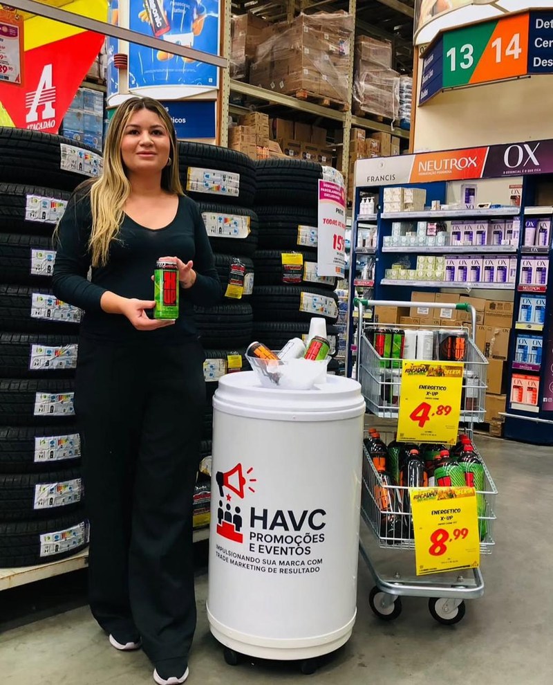 Promotora da HAVC ao lado de cooler com a marca HAVC em corredor de atacarejo, com ofertas sinalizadas