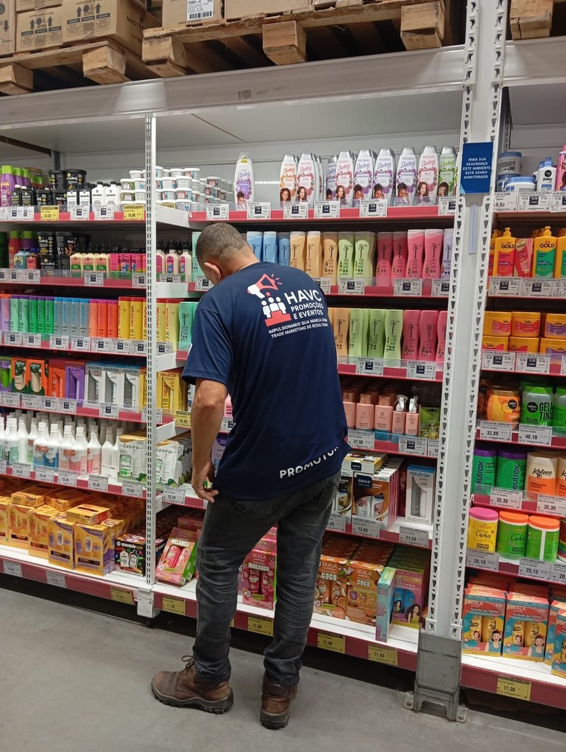 Promotor da HAVC de costas, com uniforme identificado, organizando frentes de produto na gôndola de um atacarejo